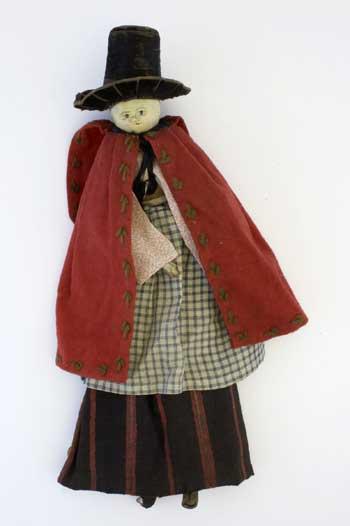 Wooden doll wearing Welsh national dress, circa 1850. Wearing a pink patterned dress, blue gingham skirt and red cloak, and a tall black hat. Separate brown and red striped skirt. Two detached legs covered in brown knitted stockings, one without a shoe.