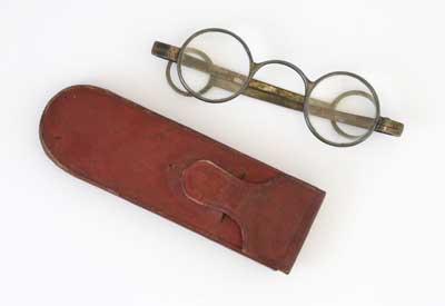 The round metal frame and the nose bridge are made of one piece. Straight shafts with a ring at the end. Flexible case with side opening. This style of glasses with arms passing over the ears was developed in the 18th Century.